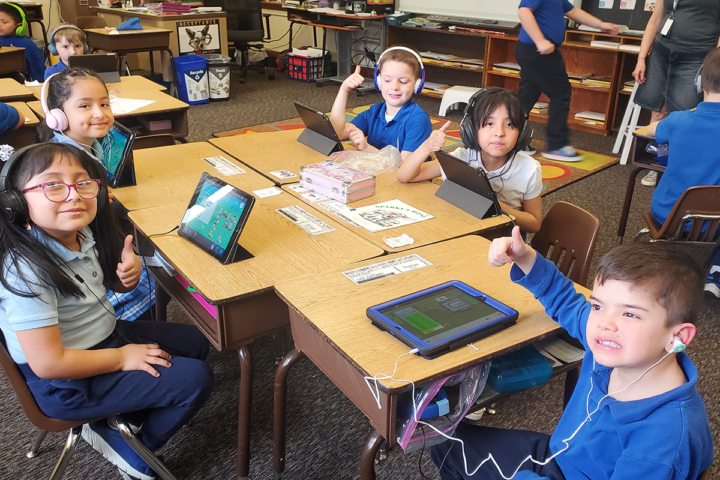 Elementary school students sit at desks with tablets and headphones.