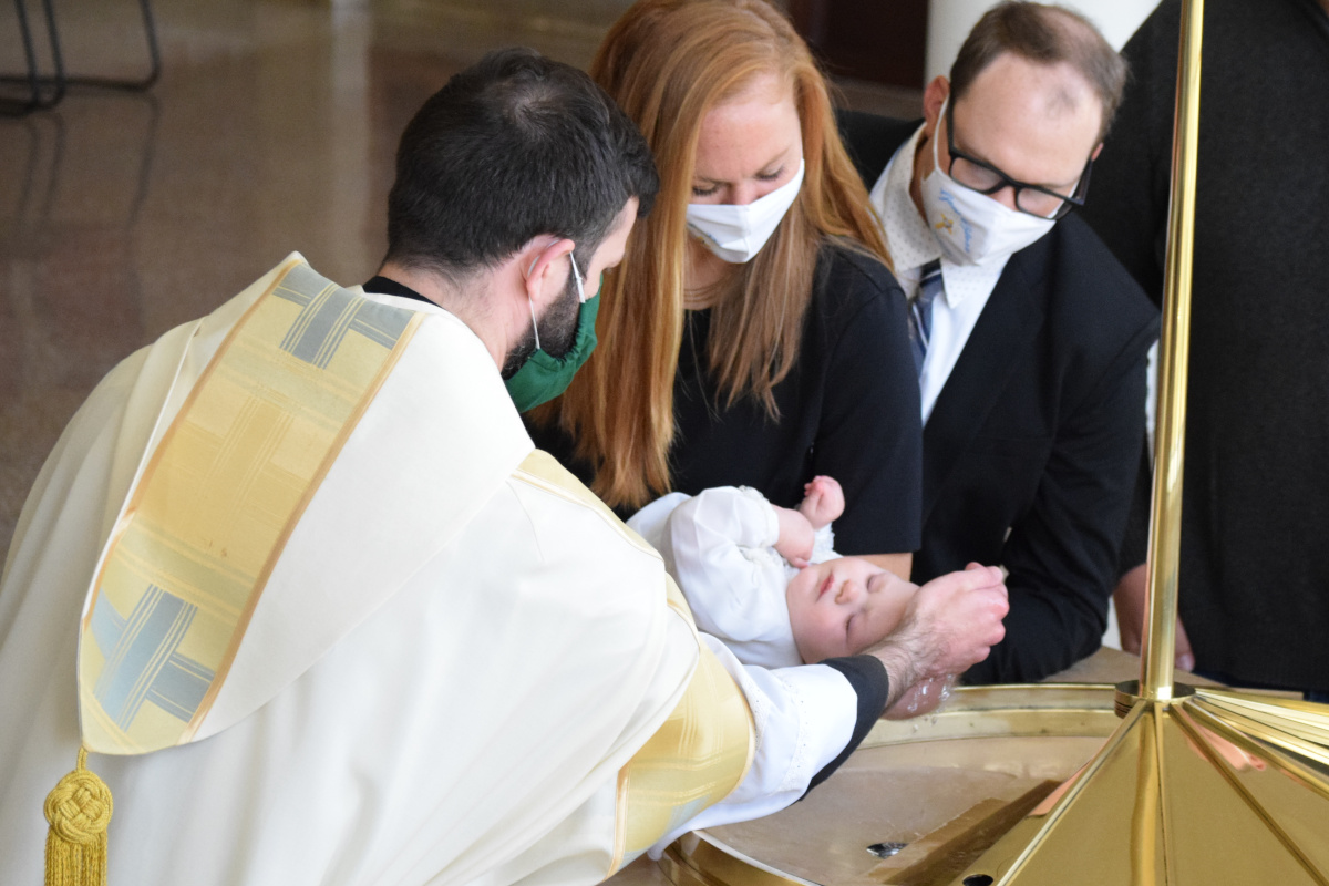 Father Joah Ellis baptizes an infant. He and the other adults are wearing face masks over their mouths and noses.