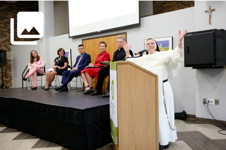 A nun in a white habit stands at a podium with her arms raised and smiling. Five people sit on a stage in the background.