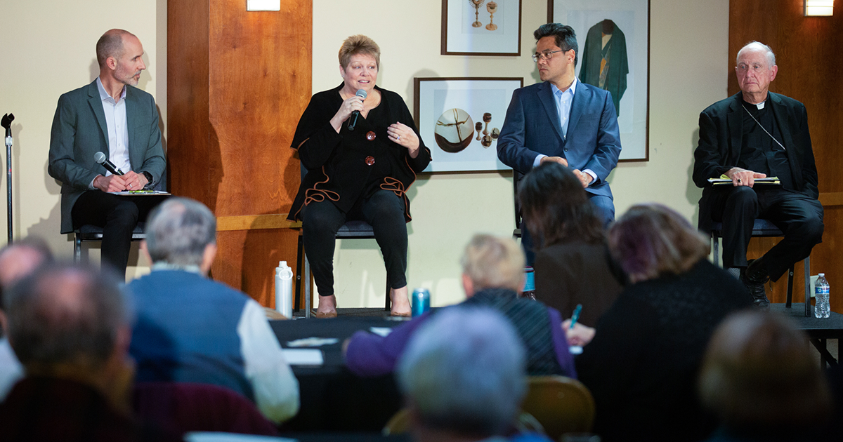 A panel of four speakers sits on a stage in front of an audience
